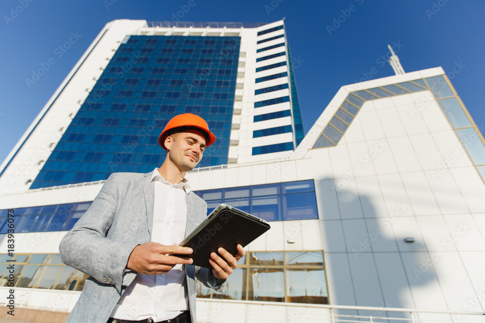 Successful business man in gray suit and protective construction orange helmet with tablet on background of building in blue white color. Engineer in helmet for workers security on office buildings. Successful business man in gray suit and protective construction orange helmet with tablet on background of building in blue white color. Engineer in helmet for workers security on office buildings.