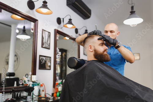 Male professional hairdresser serving client, shaving thick big beard straight razor. Ginger handsome stylish young man with short hair getting trendy haircut, black cape. Light white barber shop room
