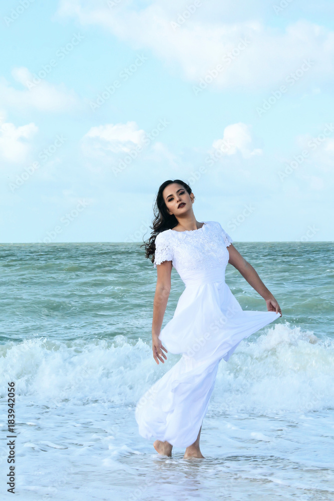 An image of attractive young woman, dressed in designer wedding, is walking on the beach with an attitude of confidence, background of blue sky, clouds, sea and sand, color, An image of attractive young woman, dressed in designer wedding, is walking on the beach with an attitude of confidence, background of blue sky, clouds, sea and sand, color,