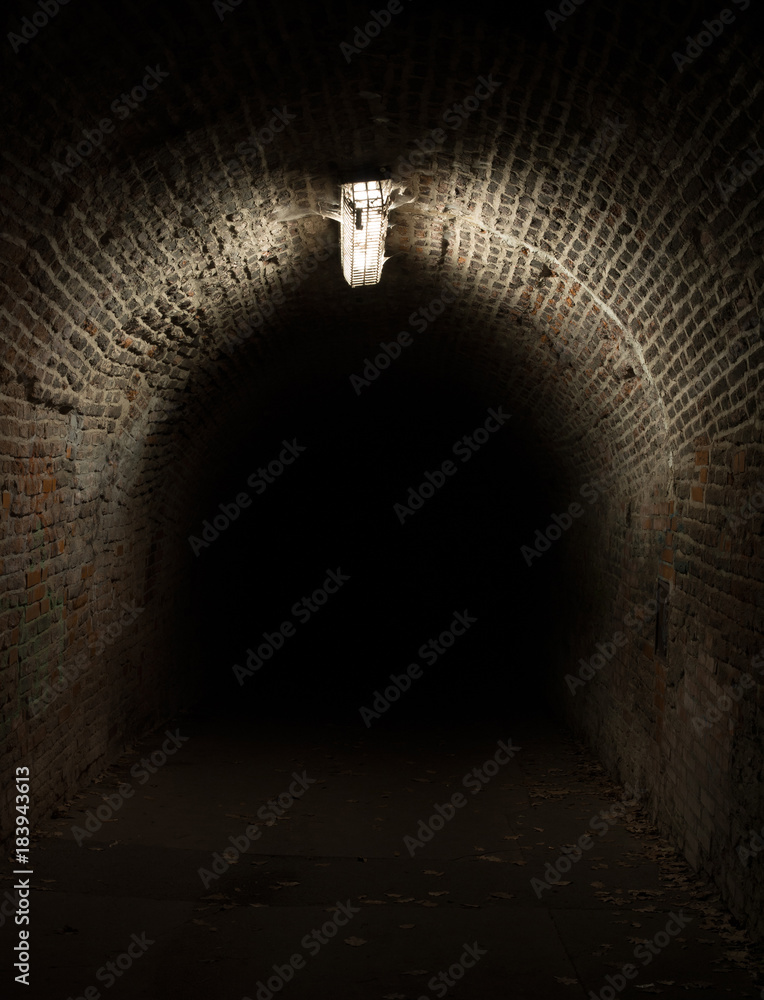 Dark tunnel and corridor during night. Light is illuminating old historical pathway made of bricks. Spooky and frightening atmosphere of dark, gloomy and murky place.