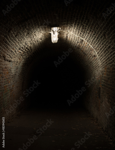 Dark tunnel and corridor during night. Light is illuminating old historical pathway made of bricks. Spooky and frightening atmosphere of dark, gloomy and murky place.