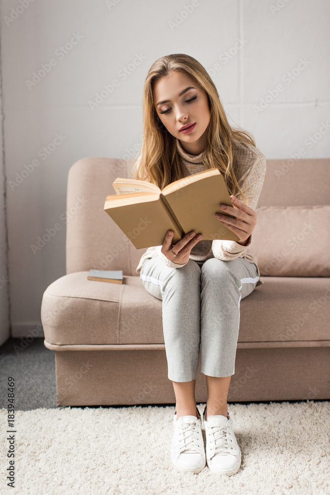 Obraz premium concentrated young woman reading book on cozy couch at home