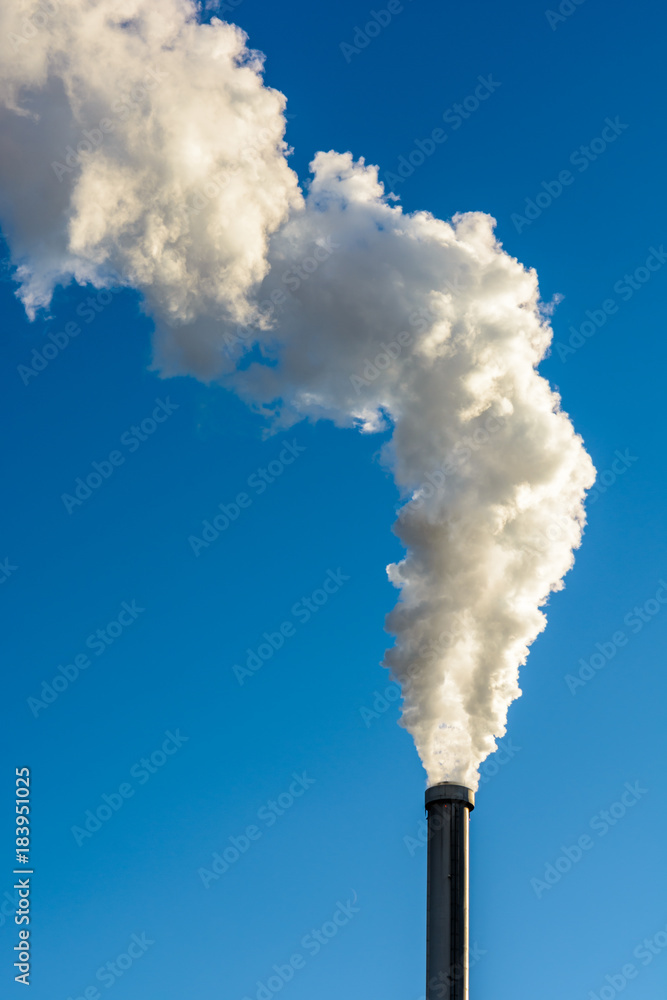 A long cloud of white smoke escaping from a metallic chimney against a deep blue sky.