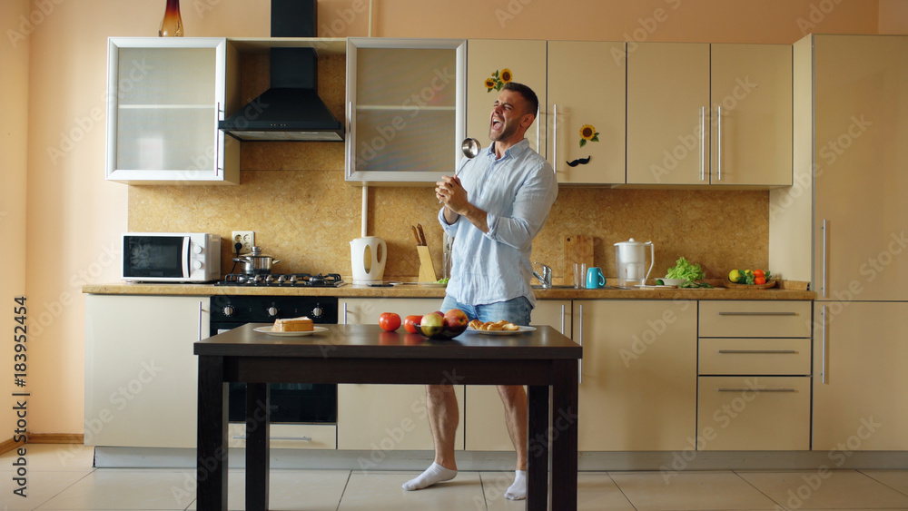 Attractive young funny man dancing and singing with ladle while cooking in the kitchen at home Attractive young funny man dancing and singing with ladle while cooking in the kitchen at home