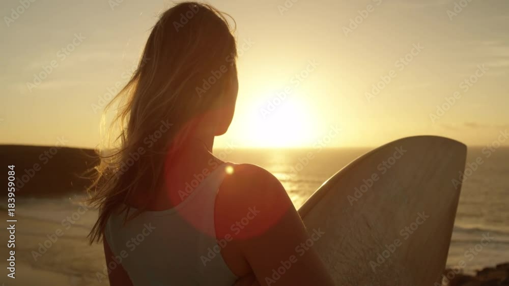 SLOW MOTION CLOSE UP: Surfer girl standing on rocky ocean cliff and watching breaking waves at golden sunset. Woman holding a surfboard, looking the waves at sunrise in Fuerteventura, Canary Islands SLOW MOTION CLOSE UP: Surfer girl standing on rocky ocean cliff and watching breaking waves at golden sunset. Woman holding a surfboard, looking the waves at sunrise in Fuerteventura, Canary Islands