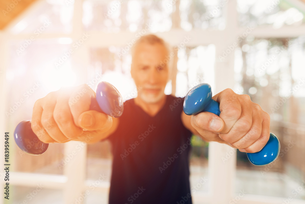 An elderly man is holding dumbbells in his hands in a nursing home. An elderly man is holding dumbbells in his hands in a nursing home.