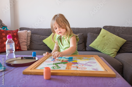 four years old blonde child with green sleeveless shirt sitting on brown sofa, playing parcheesi or parchisi or parchis, on table
 photo