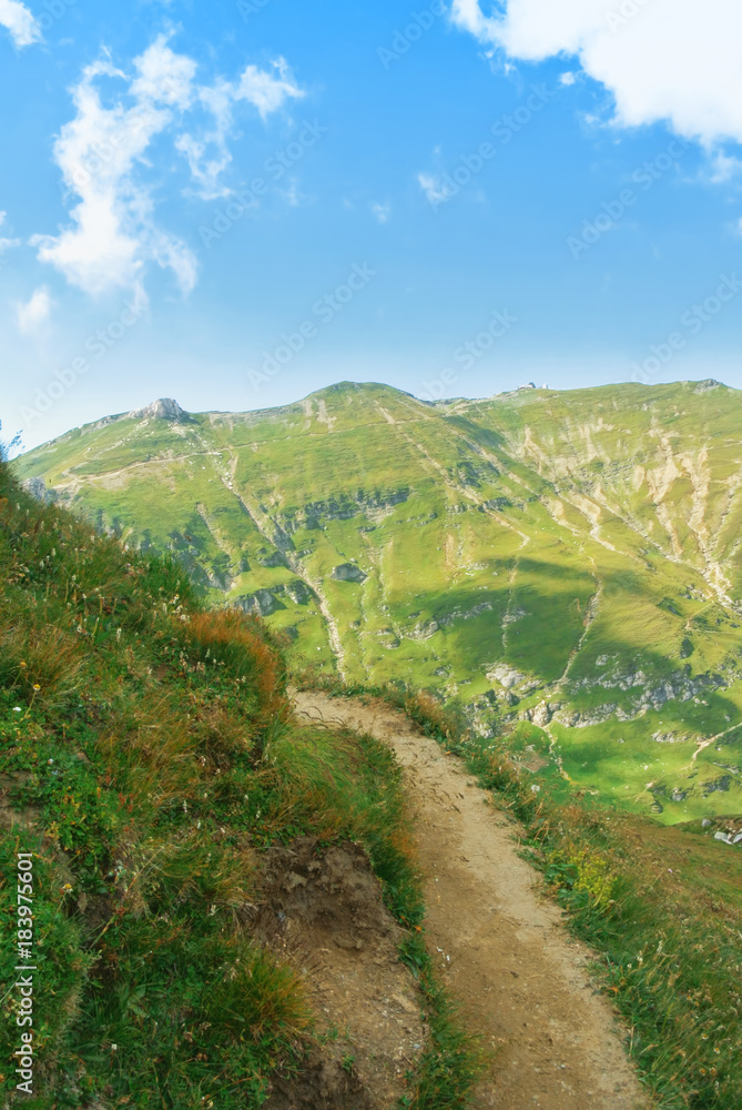 A serpentine track road narrow path at mountainside going up around a top of a hill and a beautiful panoramic view of a chain of mountains at the background on sunny summer day. Tracking in mountains.