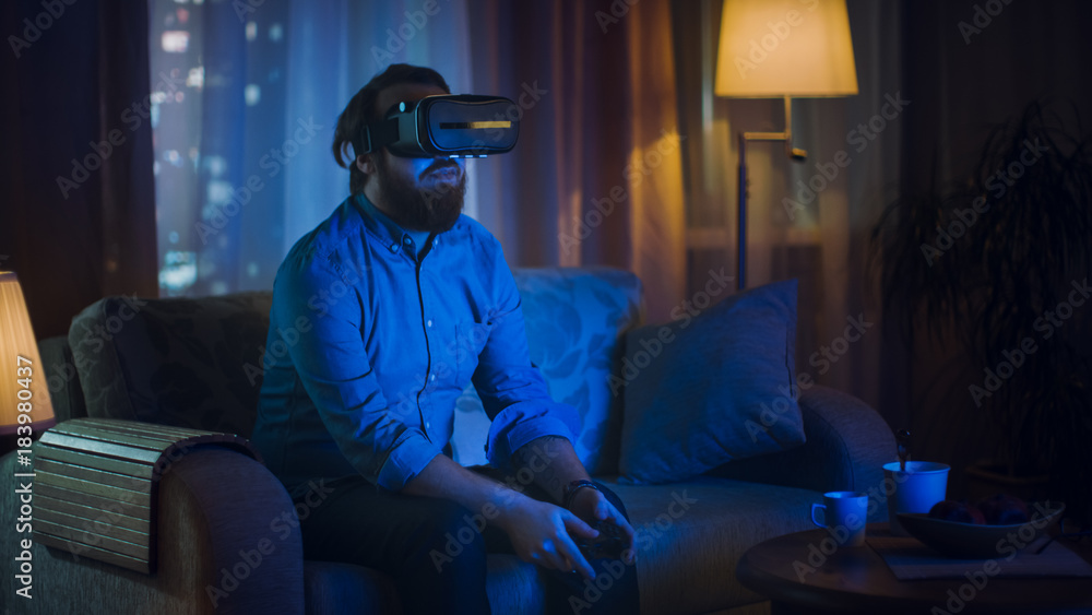 Fototapeta premium Man Sitting on a Couch in His Living Room Wearing Plays Video Games on His Console While Wearing Virtual Reality Headset.