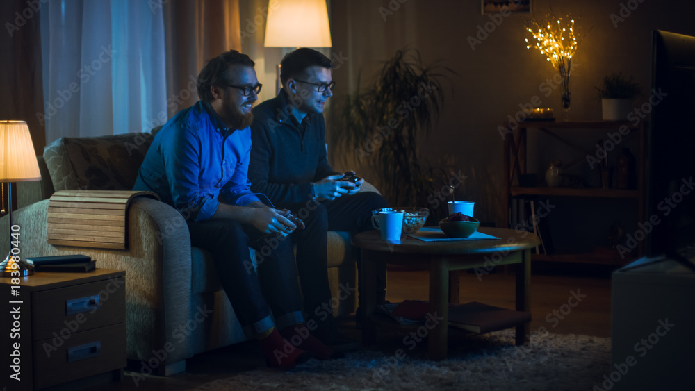 In the Evening Two Friends are Sitting on a Sofa in the Living Room and Playing Competitive Video Games. They Push and Shove Each Other Friendly. In the Evening Two Friends are Sitting on a Sofa in the Living Room and Playing Competitive Video Games. They Push and Shove Each Other Friendly.
