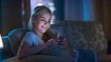 Late At Night Beautiful Woman Lying on Her Sofa in the Living Room, Using Her Smartphone and Smiling. Her Apartment Has Big City Window View.