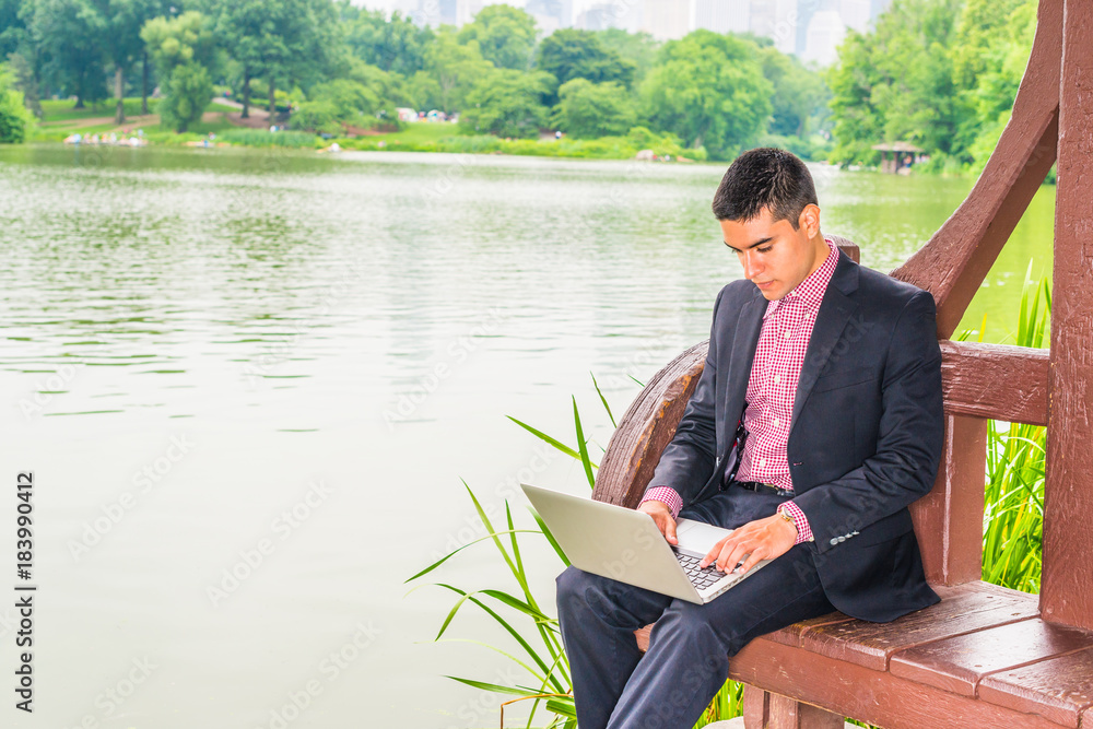 Working hard in peaceful environment. Wearing black suit, red, white patterned undershirt, a young college student sitting by lake with green trees on campus, reading, working on laptop computer.. Working hard in peaceful environment. Wearing black suit, red, white patterned undershirt, a young college student sitting by lake with green trees on campus, reading, working on laptop computer..