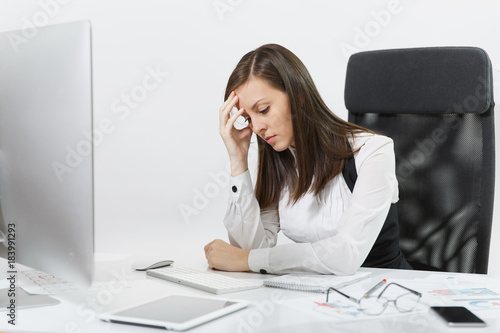 Beautiful tired perplexed and stress brown-hair business woman in suit and glasses sitting at the desk, working at contemporary computer with documents in light office on white background