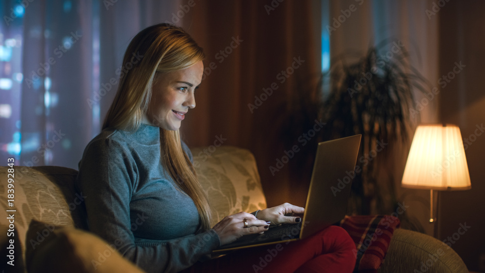 Beautiful Young Woman Works on a Laptop in Her Living Room, She is Sitting on a Sofa with Notebook on Her Lap. In the Window Big City is Seen. Beautiful Young Woman Works on a Laptop in Her Living Room, She is Sitting on a Sofa with Notebook on Her Lap. In the Window Big City is Seen.