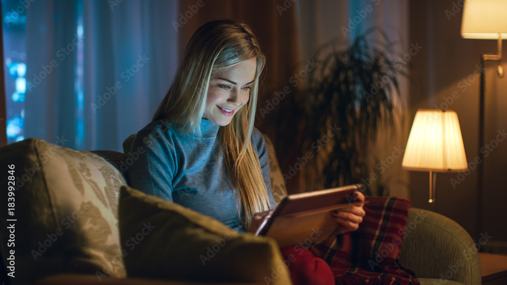 Beautiful Young Woman in Her Living Room. She is Sittin on a Sofa and Uses Tablet Computer. Behind Her Big City is Seen in the Window. - obrazy, fototapety, plakaty 