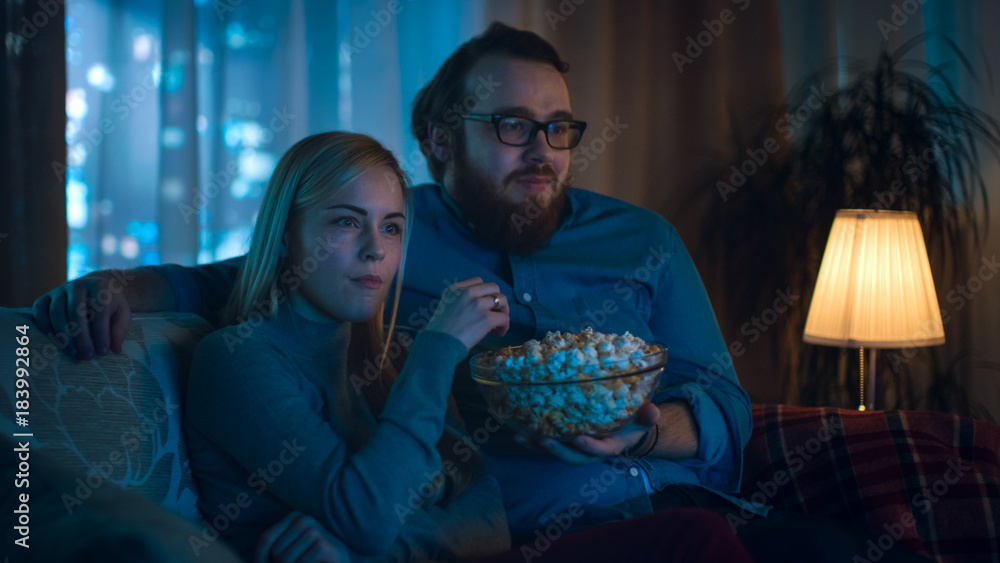 Boyfriend and Girlfriend Watching TV. They Sit on a Sofa in Their Cozy Living Room and Eat Popcorn. It's Evening.