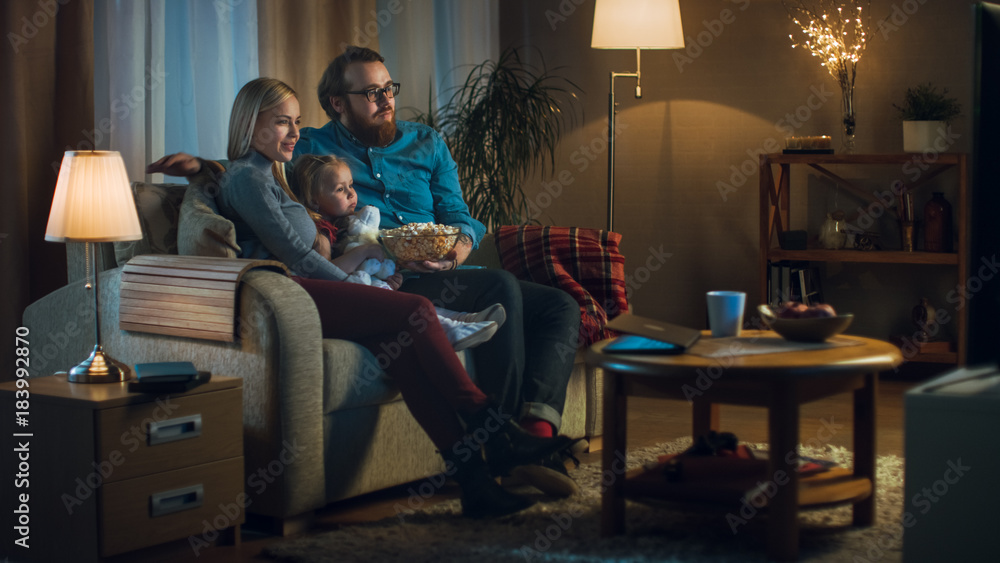 Long Shot of a Father, Mother and Little Girl Watching TV. They Sit on a Sofa in Their Cozy Living Room and Eat Popcorn. It's Evening. Long Shot of a Father, Mother and Little Girl Watching TV. They Sit on a Sofa in Their Cozy Living Room and Eat Popcorn. It's Evening.