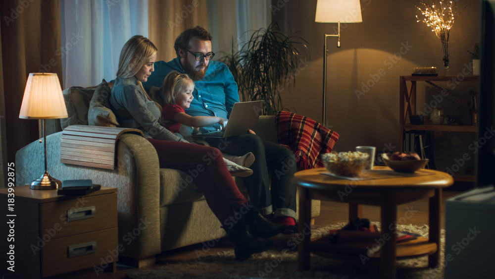 Long Shot of a Father, Mother and Little Girl Watching TV. They Sit on a Sofa in Their Cozy Living Room, Father Holds Laptop on His Knees. It's Evening. Long Shot of a Father, Mother and Little Girl Watching TV. They Sit on a Sofa in Their Cozy Living Room, Father Holds Laptop on His Knees. It's Evening.