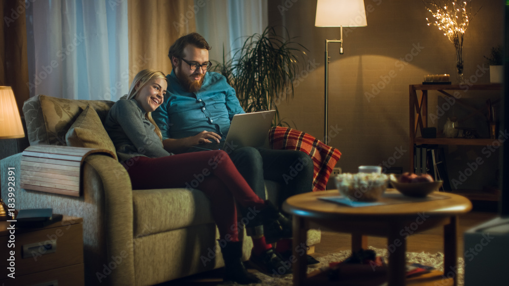 Girlfriend and Boyfriend Sitting on a Sofa with Laptop on His Knees, Watching Videos and Having Fun. Living Room is Cozy. It's Evening. Girlfriend and Boyfriend Sitting on a Sofa with Laptop on His Knees, Watching Videos and Having Fun. Living Room is Cozy. It's Evening.