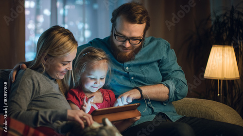 Father, Mother and Little Daughter Reading Children's Book on a Sofa in the Living Room. It's Evening.