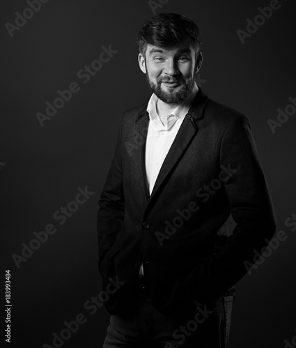 Emotion enjoying handsome beard male posing in fashion suit and white style shirt looking on dark grey shadow background. Black and white portrait