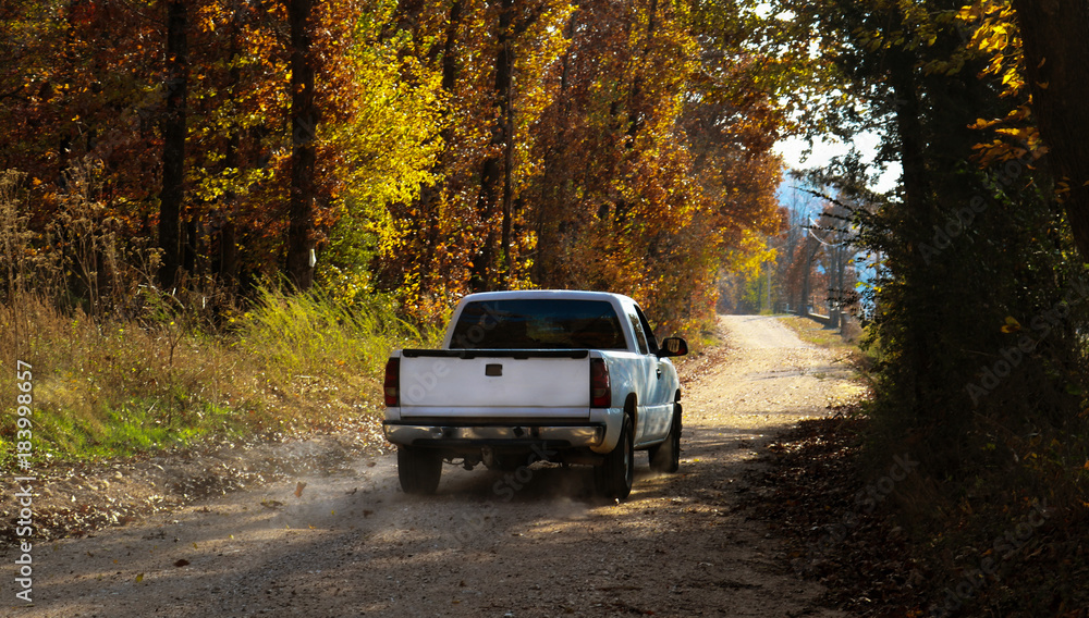White pickup truck driving down dusty dirt road with fall leaves and dust behind - obrazy, fototapety, plakaty 