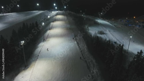 Aerial view: Skiers and snowboarders going down the slope in winter night. Skiers and snowboarders enjoying on slopes of ski resort in winter season. 4K video, aerial footage.