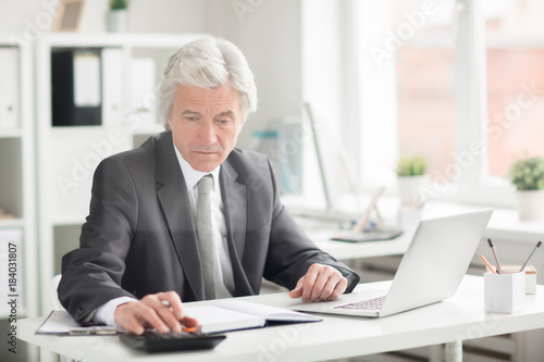 Experienced economist in suit sitting by desk in office and making calculations