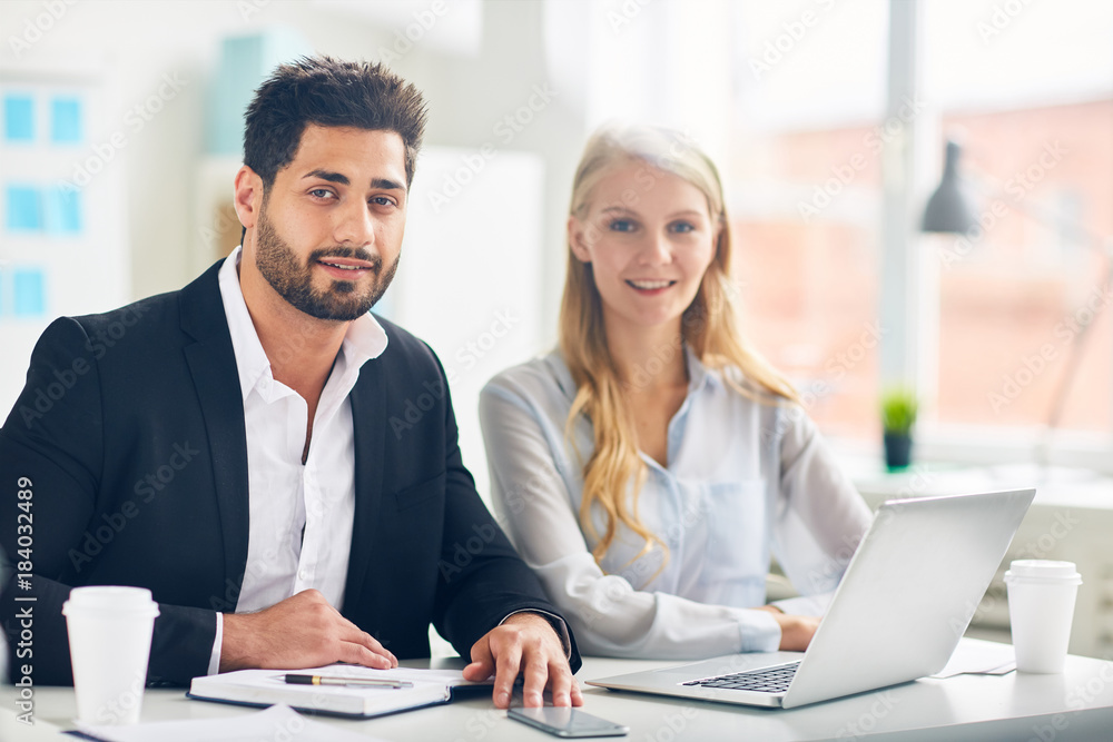 Two successful colleagues in formalwear looking at camera by their workplace in office Two successful colleagues in formalwear looking at camera by their workplace in office