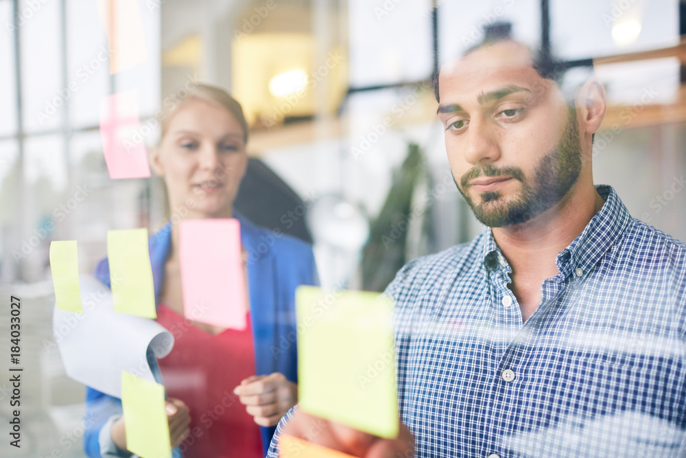 Obraz premium Businessman looking at one of notepapers on transparent office board with colleague near by