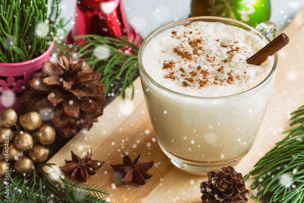 Eggnog, traditional drink for Christmas celebration party in glass topped with ground nutmeg and cinnamon stick, top view with copy space in Xmas theme decoration and snowfall effect background. Eggnog, traditional drink for Christmas celebration party in glass topped with ground nutmeg and cinnamon stick, top view with copy space in Xmas theme decoration and snowfall effect background.