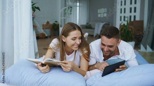 Handsome young man using tablet computer show photos on tablet computer to his girfriend reading book and lying in bed at home photo