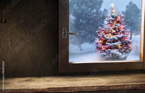 Christmas background. A cup of tea  books  a candle and a knitted scarf on the window background. Outside the window is winter  snowfall and Christmas tree lights. Cozy warm home holiday atmosphere.