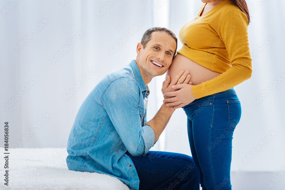 Fototapeta premium I hear him. Positive delighted future father keeping smile on his face and putting hands on the baby belly while looking forward