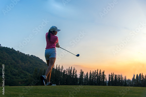 Asian woman golf player doing golf swing tee off on the green sunset evening time, she presumably does exercise. Healthy and Lifestyle Concept.