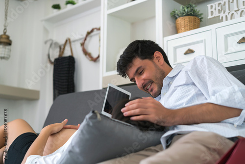 Handsome husband reading book and asian pregnant woman playing tablet computer with lying on sofa in the living room at home. concept of happy family takecare and healthy.