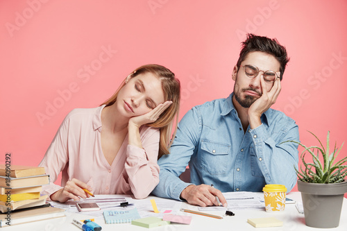 Portrait tired crew of office workers sit at table, fall asleep after working long hours on preparing startup project, feel tiredness, isolated over pink background. People and overworking concept photo