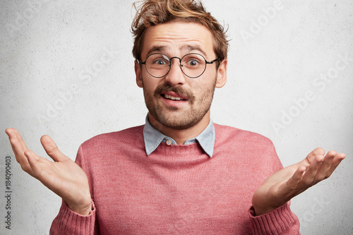 Clueless doubtful male wonk wears round spectacles, shrugs shoulders in bewilderment as doesn`t know answer on question, hesitates, isolated over white concrete background. Body language concept photo