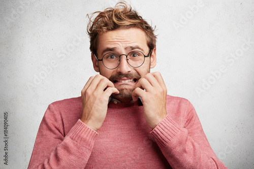 Unhappy worried sad man clenches teeth, bites nails, worries to hear results of test, has sorrorful mournful expression, regrets about something, isolated over white concrete wall. Gloomy unhappy male photo