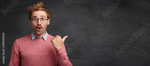 Horizontal shot of shocked male student with stylish hairdo, stares through spectacles, being surprised to indicate something at copy space for your promotional text or hearder. Advertising concept photo