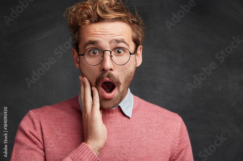 Close up portrait of shocked man looks with terrified expression, being in stupor, keeps jaw dropped out, puzzled to find out about expulsion or dismissal, isolated over black chalk background