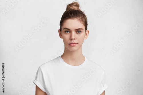 Serious pleasant looking woman with dark blonde hair tied in bun, wears casual white t shirt going to have walk outdoor with pet, has healthy pure skin. Pretty young female model poses in studio