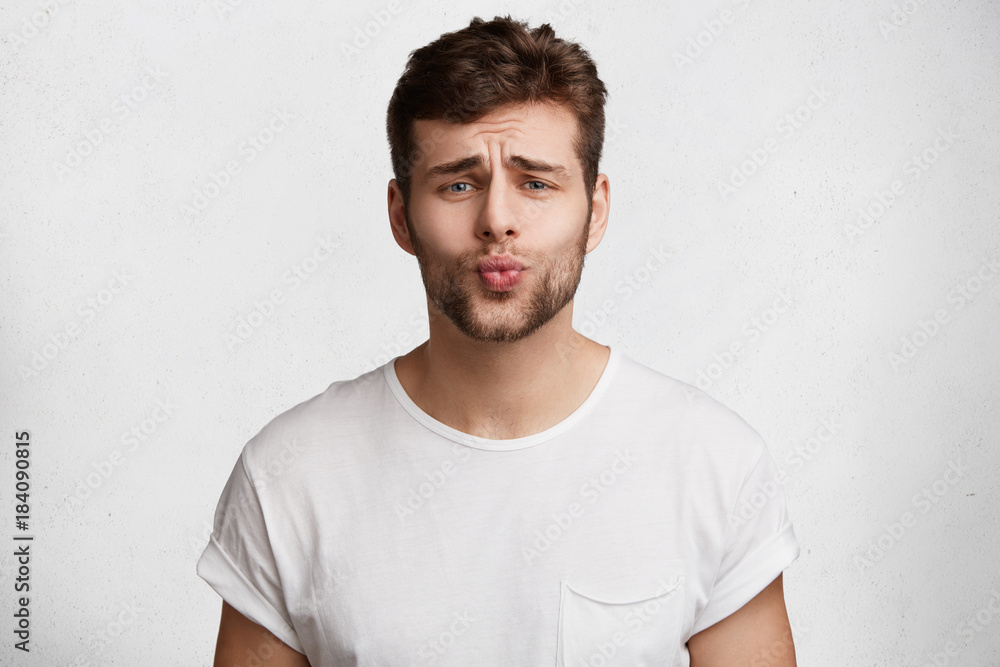 Horizontal portrait of bearded stylish young male rounds lips and has pleading expression, asks for forgiveness in her girlfriend as comes home late, isolated over white concrete background. Horizontal portrait of bearded stylish young male rounds lips and has pleading expression, asks for forgiveness in her girlfriend as comes home late, isolated over white concrete background.
