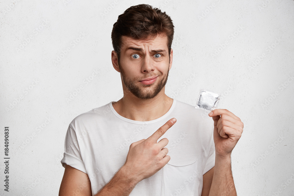 Displeased handsome man with stubble wears casual white t shirt, holds rubber condom in hands, advertizes its quality. Pleasant looking unshaven stylish young male being against of contraceptives Displeased handsome man with stubble wears casual white t shirt, holds rubber condom in hands, advertizes its quality. Pleasant looking unshaven stylish young male being against of contraceptives