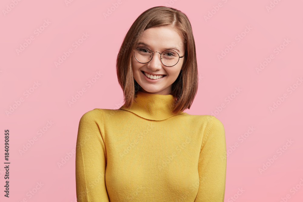 Positive pleasant looking young female model with short dark hair, wears yellow poloneck sweater, smiles pleasantly at camera as being glad to recieve good news, isolated over pink background Positive pleasant looking young female model with short dark hair, wears yellow poloneck sweater, smiles pleasantly at camera as being glad to recieve good news, isolated over pink background