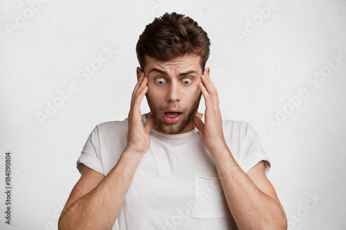 Φωτογραφία Terrified shocked male looks with bugged eyes down, sees something awful, can`t believe his eyes, being in stupor, keeps hands on temples, isolated over white studio background