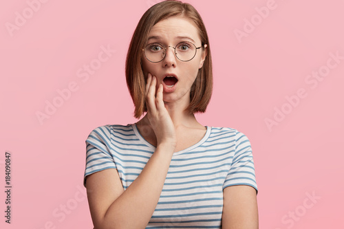 Surprised young female model with short hair, wears round optical glasses and striped t shirt, looks with terror at camera as notices something unexpected or hears bad news, isolated over pink wall