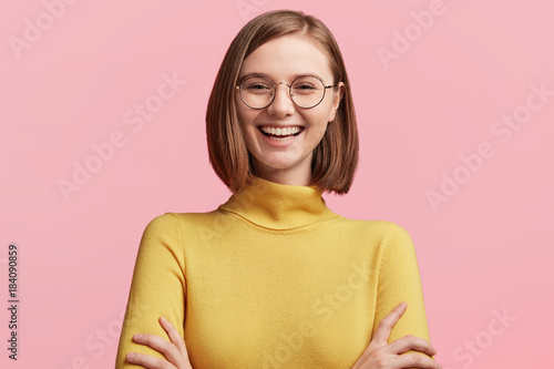 Cheerful female student has fun during break in colleague, laughes at jokes of groupmates, keeps hands crossed, wears round optical glasses and yellow turtleneck sweater. Positive emotions concept
