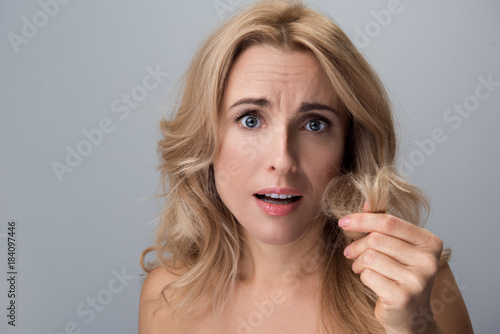 Portrait of disappointed middle-aged woman with naked shoulders is standing and holding damaged ends of her hair in hand. She is looking at camera unhappily. Isolated background. Haircare concept
