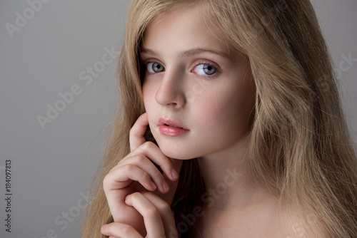 Close-up of face of pensive cute little girl with long hair is looking at camera joylessly while standing and touching her face. Isolated background